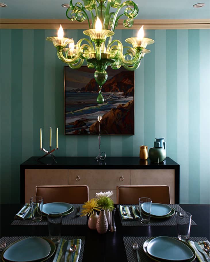 Dining room with green chandelier, striped walls, set table, and landscape painting above a sideboard.