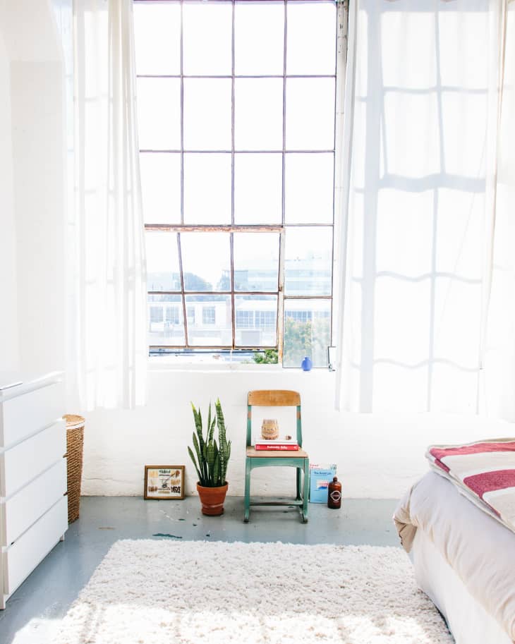 Bright bedroom with large window, green chair, potted plant, white rug, and bed with pink blanket.