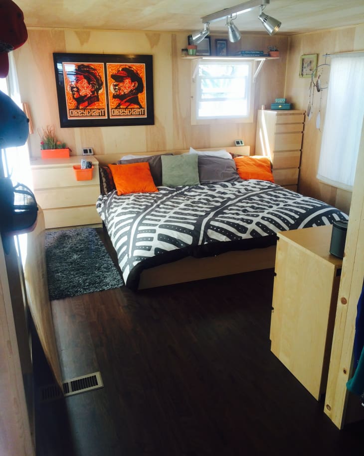 Bedroom with a large bed, geometric duvet, orange and gray pillows, wooden furniture, and two vibrant wall posters.