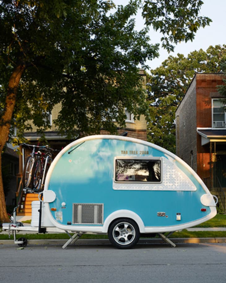 Blue teardrop camper trailer parked on a residential street with trees and houses in the background.