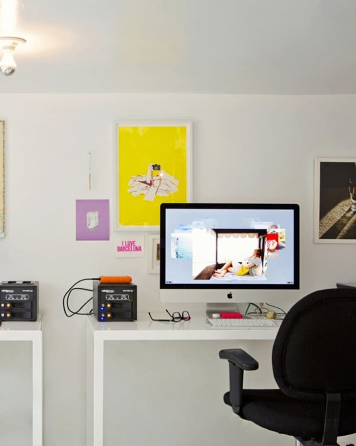 Home office with a computer on a white desk, black chair, colorful wall art, and electronic equipment.