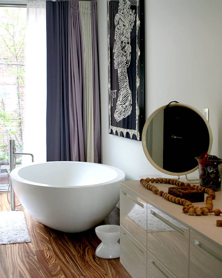 Modern bathroom with a round freestanding tub, wood floor, round mirror, and abstract wall art.