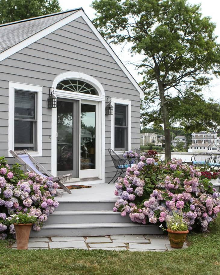 Small gray cottage with hydrangeas, patio chairs, and a view of the lake.