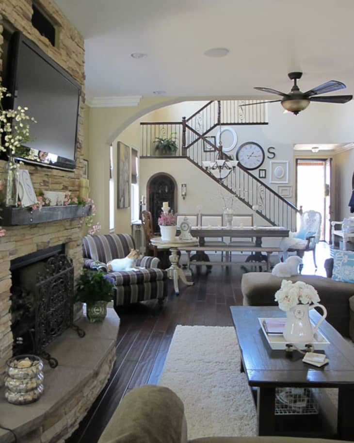 Living room with stone fireplace, striped sofa, wooden coffee table, and staircase with wall clocks and decor.