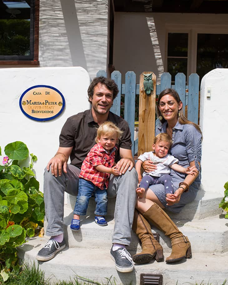 Family sitting on front steps of a house with a blue gate and a welcome sign, surrounded by greenery.