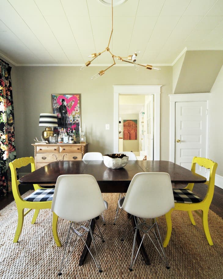 Dining room with dark wood table, white and yellow chairs, floral curtains, and a modern gold chandelier.
