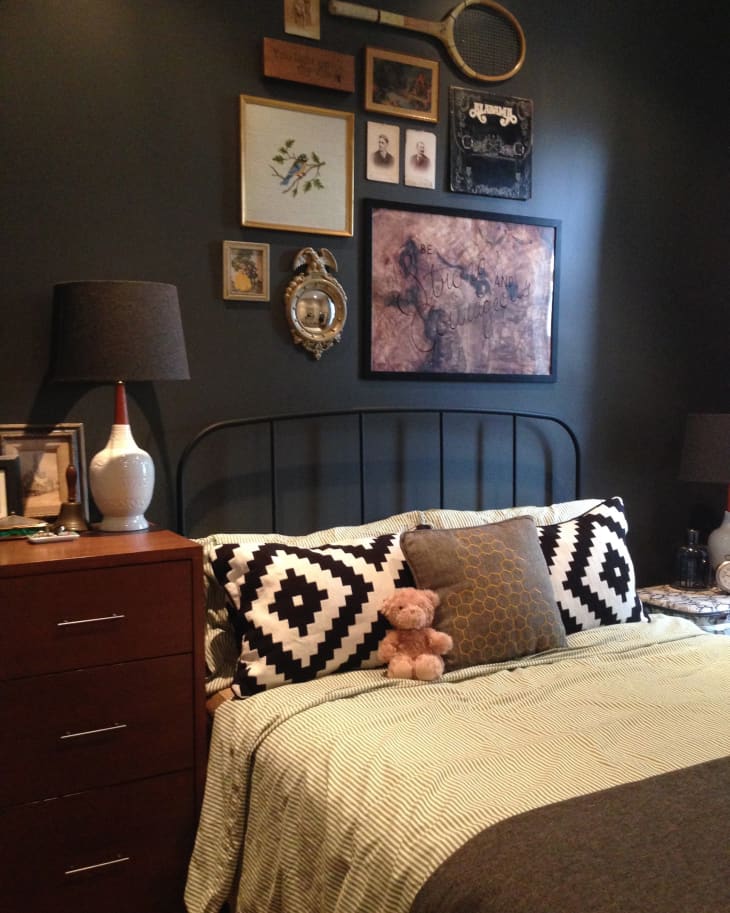 Bedroom with dark walls, eclectic wall art, black and white pillows, a teddy bear, and a wooden dresser with a lamp.