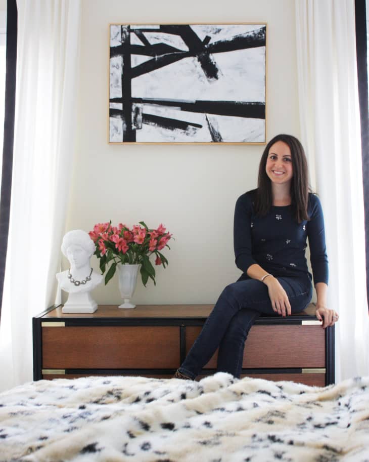 Woman sitting on a dresser with a black and white abstract painting, flowers, and a bust in a bedroom.