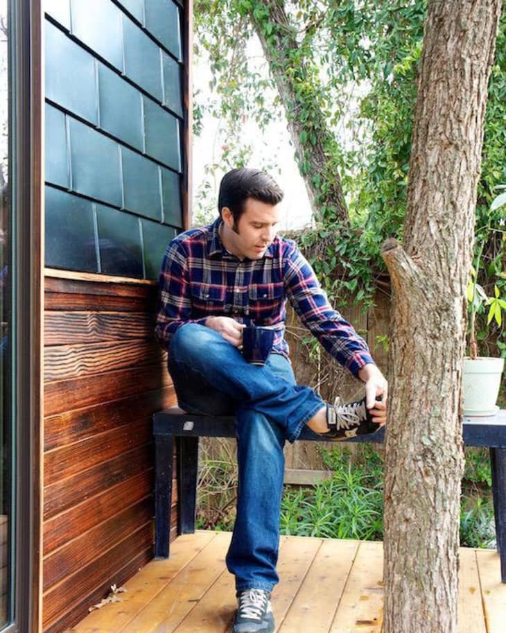 Man in plaid shirt sitting on a porch bench, holding a mug, adjusting shoe, surrounded by trees.