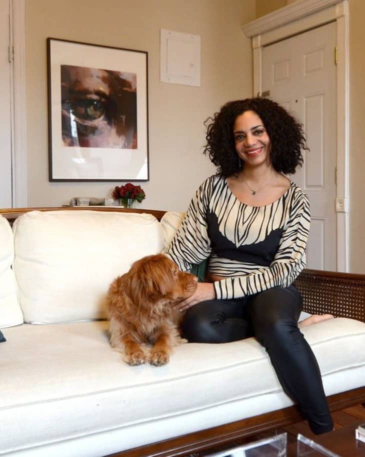 Woman sitting on a cream sofa with a brown dog, abstract eye artwork on wall, and a small vase of flowers nearby.