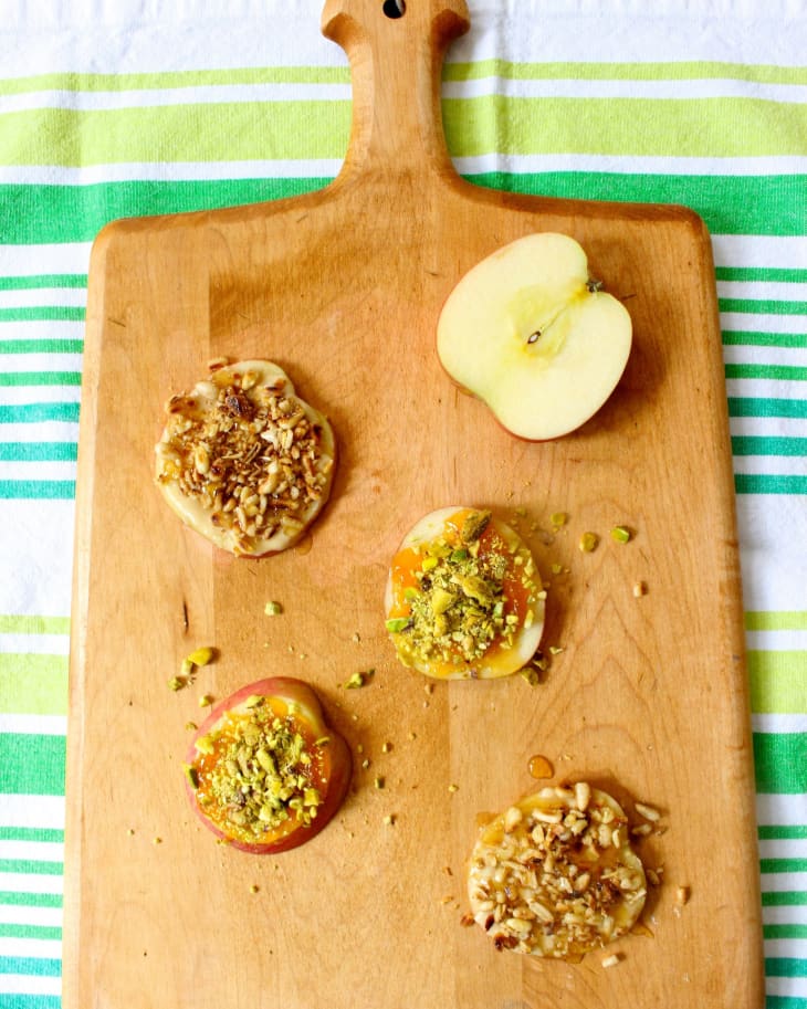 Apple slices topped with nuts and seeds on a wooden cutting board, next to a halved apple.