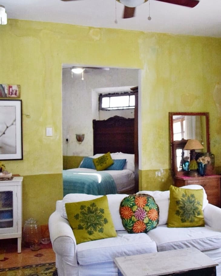 Cozy living room with white sofa, colorful pillows, yellow walls, and view into a bedroom with teal bedding.