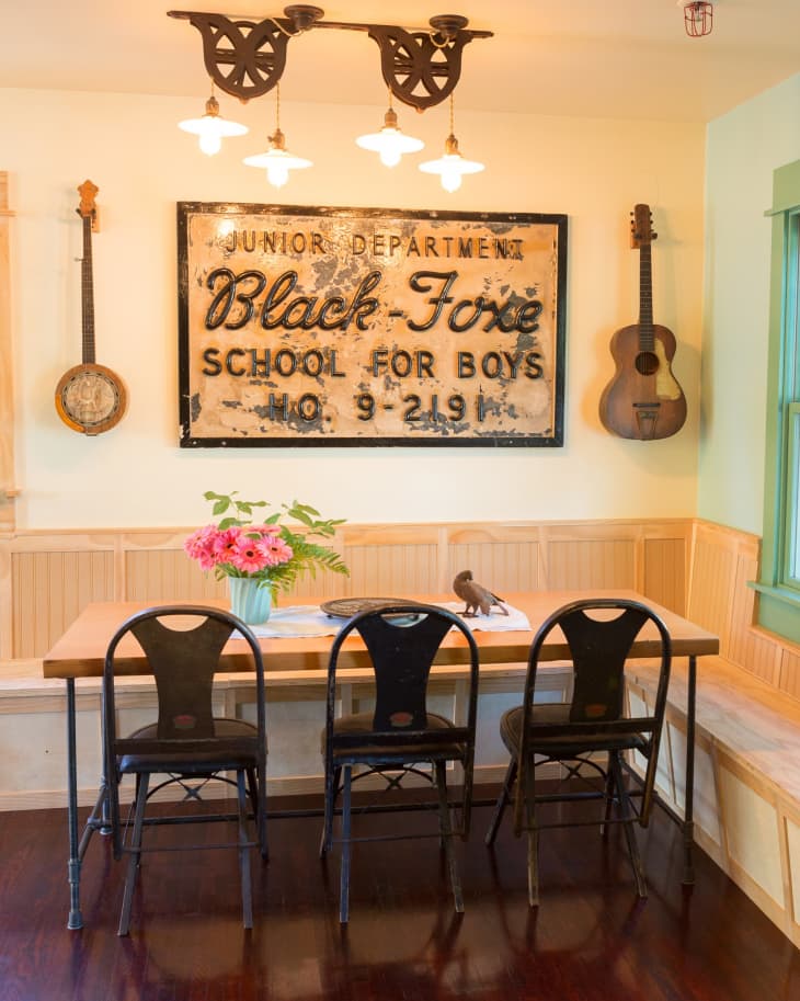Dining area with vintage sign, wooden table, metal chairs, banjo, guitar, and pink flowers in a vase.