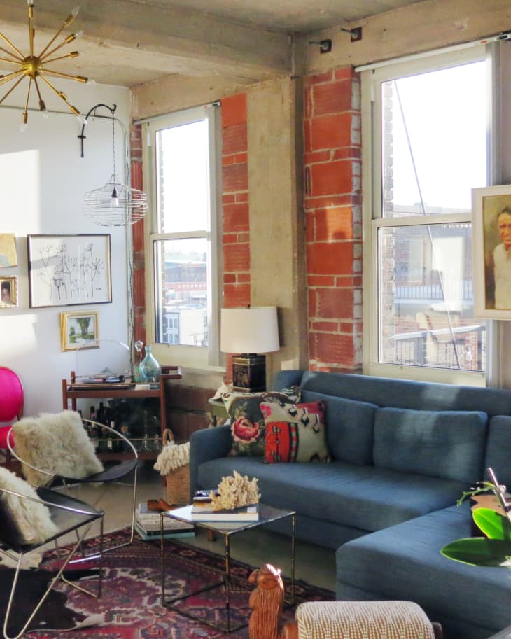 Loft living room with blue sofa, colorful pillows, glass coffee table, eclectic decor, and large windows.
