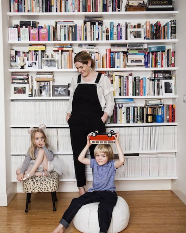 Woman in black overalls with two children in front of a bookshelf; one child holds a toy bus.