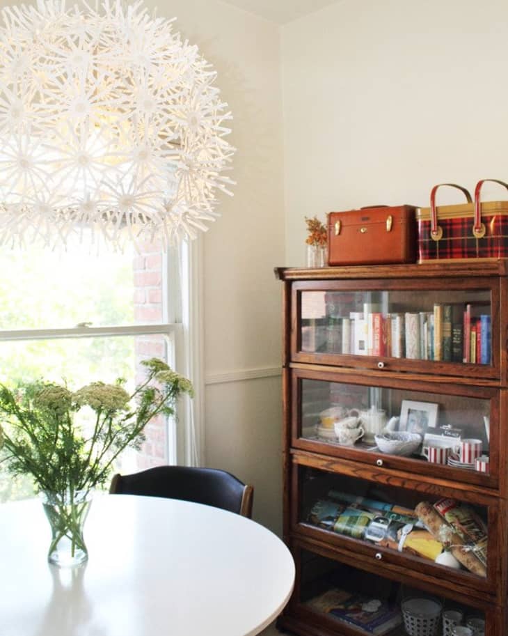 Dining area with round white table, floral centerpiece, wooden cabinet with books and dishes, and a decorative pendant light.