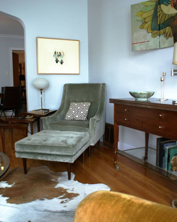 Velvet armchair with geometric pillow, wooden sideboard, abstract art, and cowhide rug in a cozy living room.