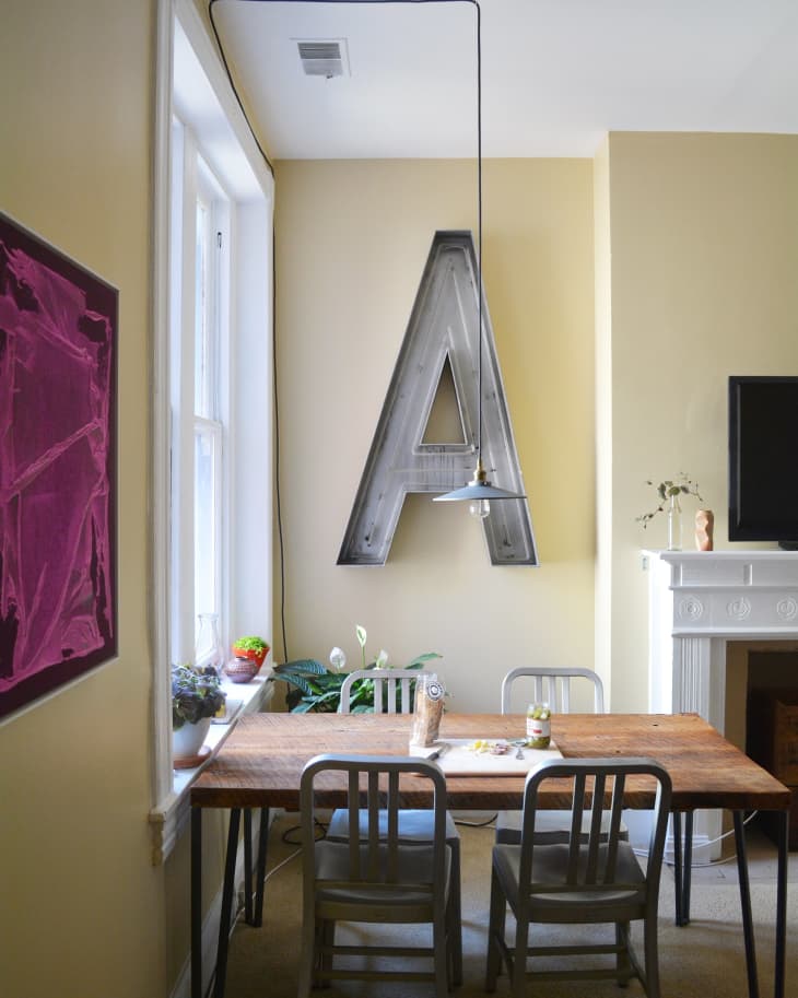 Dining area with wooden table, metal chairs, large letter "A" wall decor, and potted plants by the window.