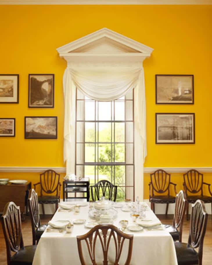 Elegant dining room with yellow walls, white tablecloth, classic wooden chairs, and framed art around a large window.