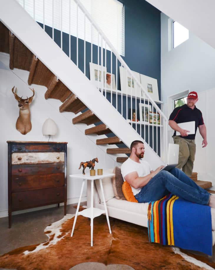Man sitting on a sofa under stairs with a colorful blanket, a small table, and a chest of drawers with a deer head mounted above.