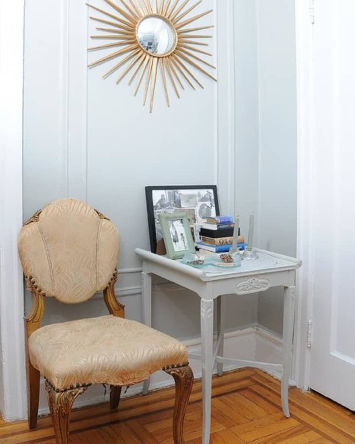 Elegant corner with a vintage chair, ornate side table, sunburst mirror, and framed photos on a wooden floor.