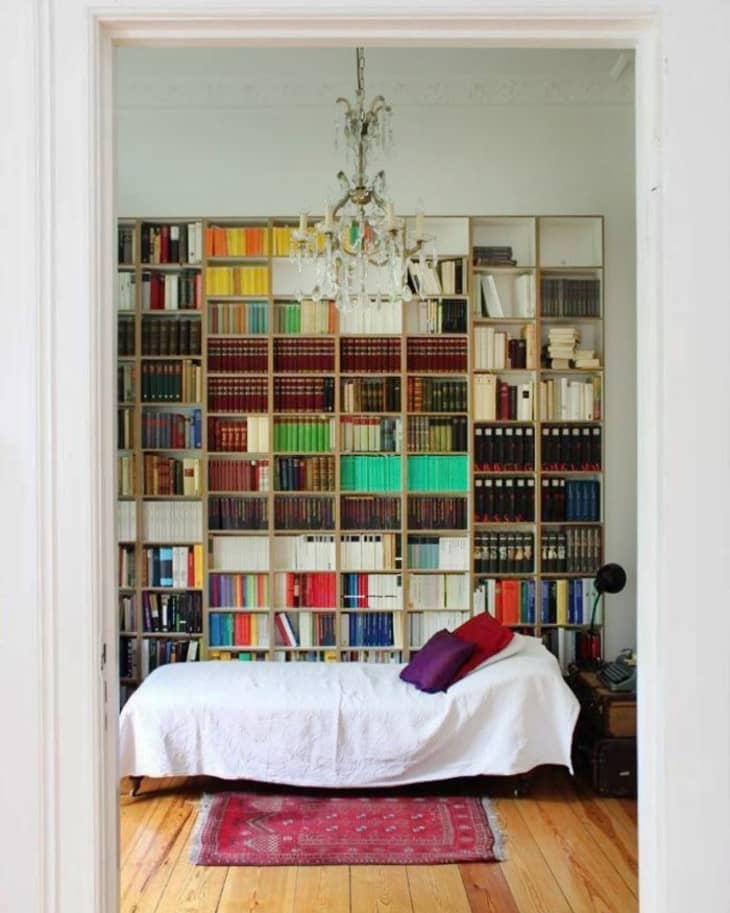 Room with a large bookshelf, white daybed, colorful books, red rug, and a chandelier.