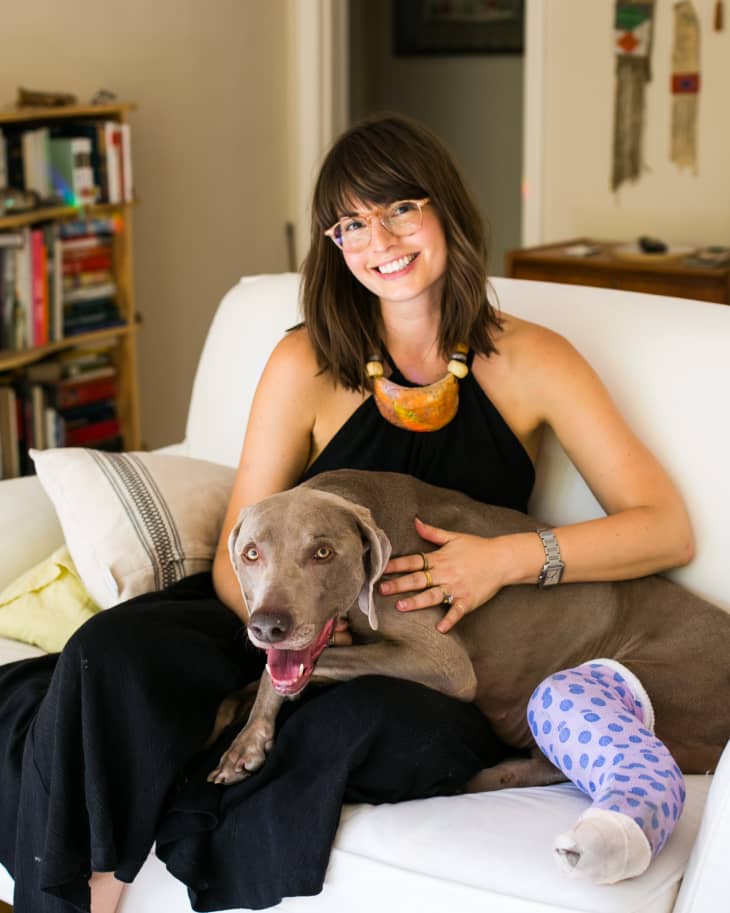 Woman sitting on a white sofa with a gray dog wearing a purple polka dot cast, bookshelf in the background.