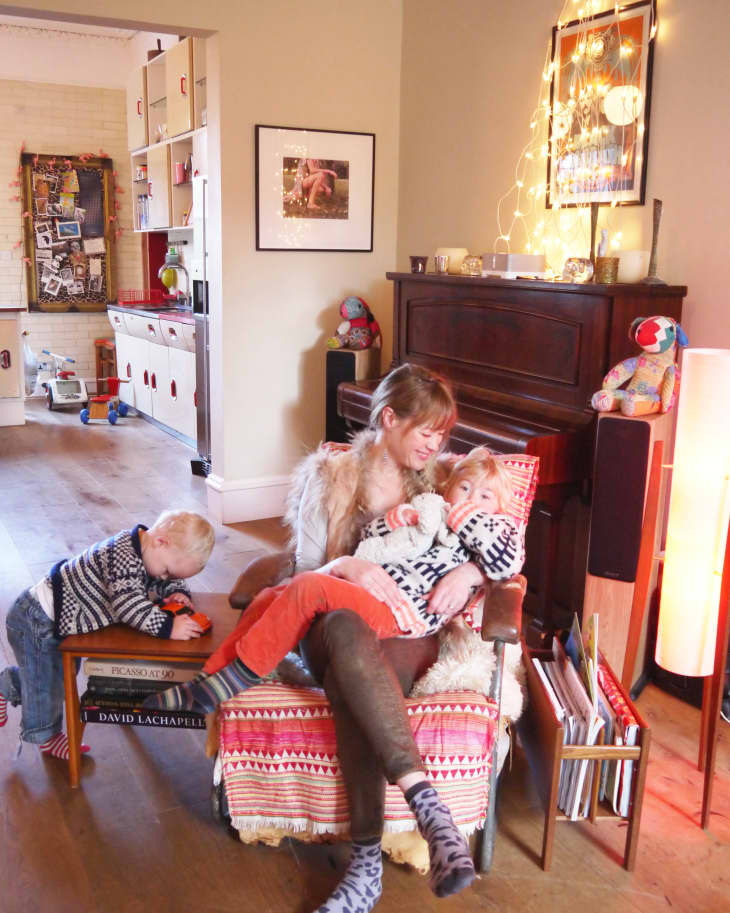 Cozy living room with a woman and child on a patterned chair, another child playing nearby, and a piano adorned with string lights.