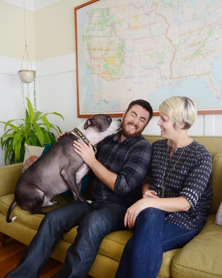 Couple sitting on a green sofa with a dog, large map on wall, and hanging plant.