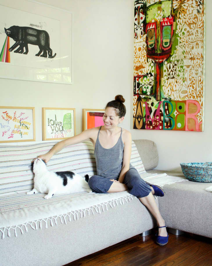 Woman petting a black and white cat on a striped sofa, surrounded by colorful artwork on the walls.