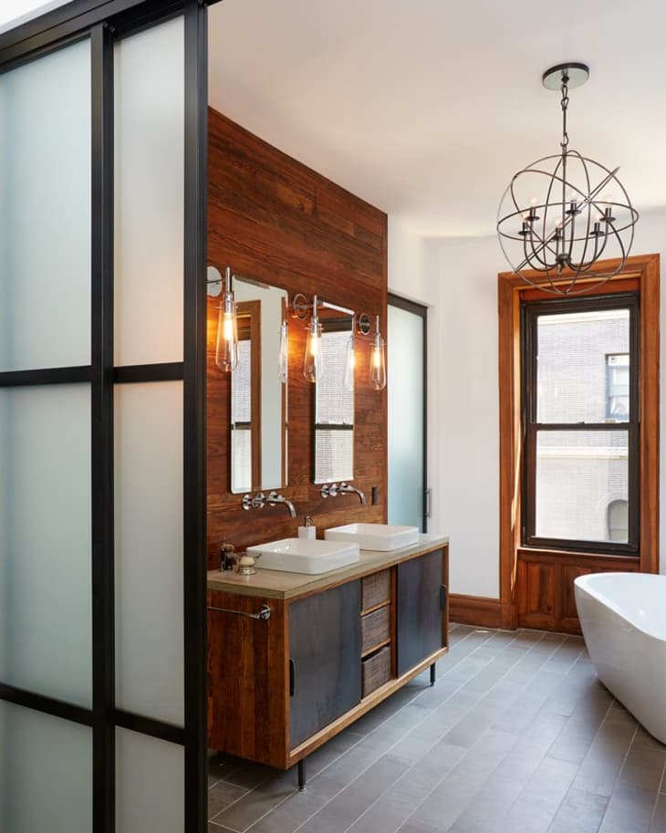 Modern bathroom with wood accents, double sinks, pendant lights, and a freestanding tub near a large window.