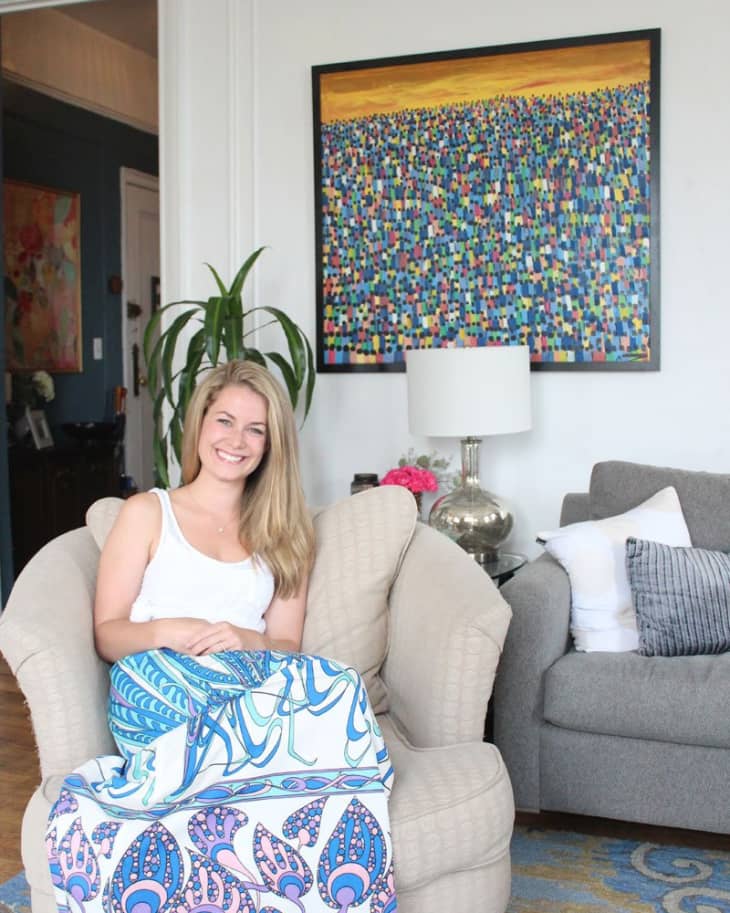Woman sitting on a beige armchair with a colorful blanket, next to a gray sofa and abstract painting.