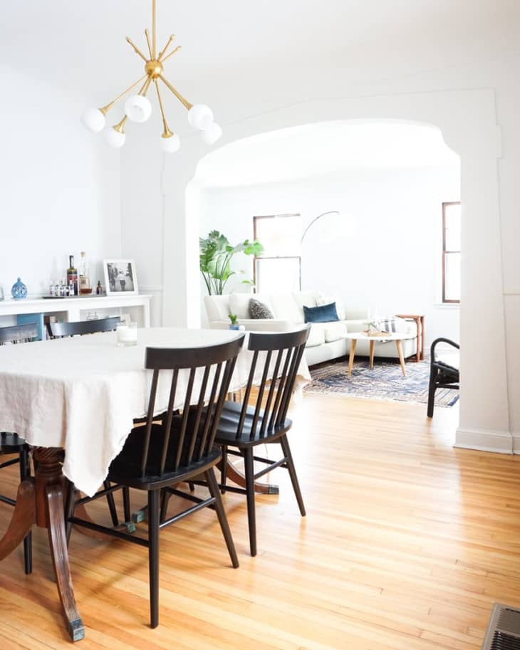 Dining room with black chairs, white tablecloth, gold chandelier, and view into a bright living room with a white sofa.