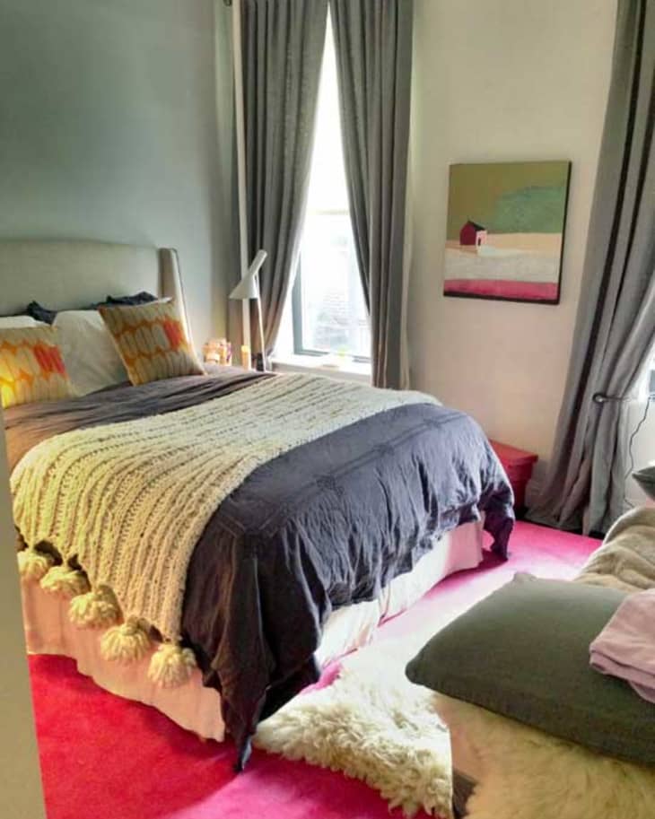 Cozy bedroom with a gray bedspread, knit throw, colorful pillows, gray curtains, and a painting on the wall.