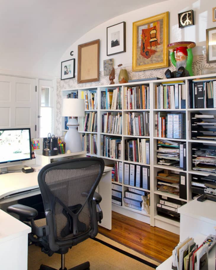 Home office with a black ergonomic chair, white desk, computer monitor, bookshelves filled with books, and framed art.