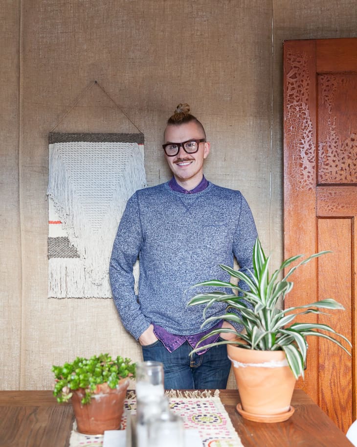 Person in a blue sweater standing by a wooden table with potted plants and a woven wall hanging.