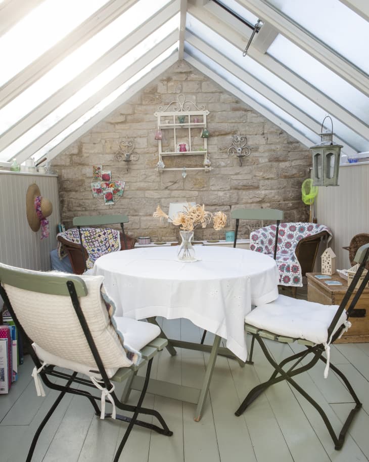 Cozy sunroom with a round table, folding chairs, floral cushions, and a stone wall with decorative shelves and lanterns.