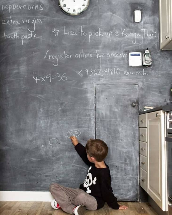 Child drawing on a chalkboard wall in a kitchen with notes and a clock.