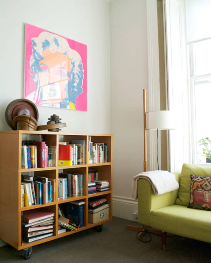 Living room with green sofa, wooden bookshelf, colorful artwork, and floor lamp by a large window.