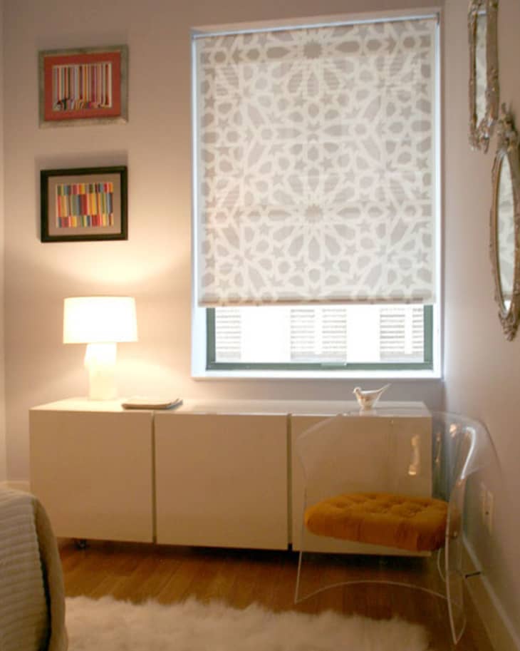 Cozy room with patterned window shade, white sideboard, lamp, clear chair with yellow cushion, and framed art.
