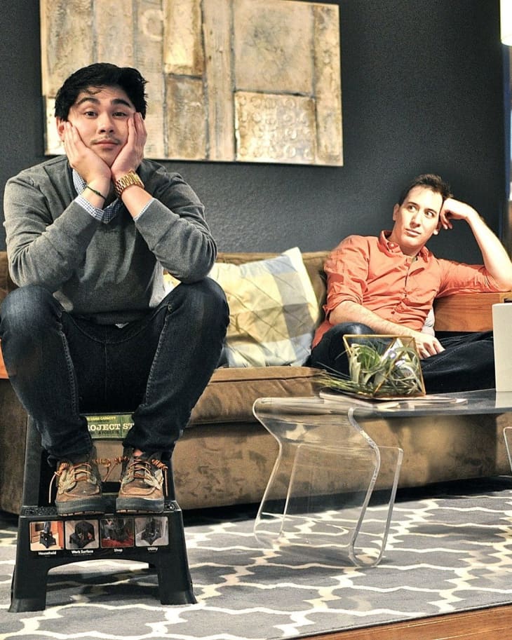 Two men in a living room, one sitting on a stool, the other on a sofa, with a geometric rug and abstract wall art.