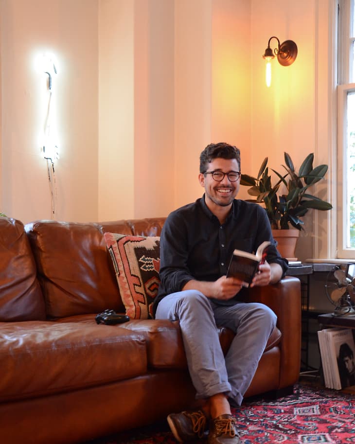 Man sitting on a brown leather sofa, holding a book, with a patterned rug, potted plant, and wall sconce in the background.