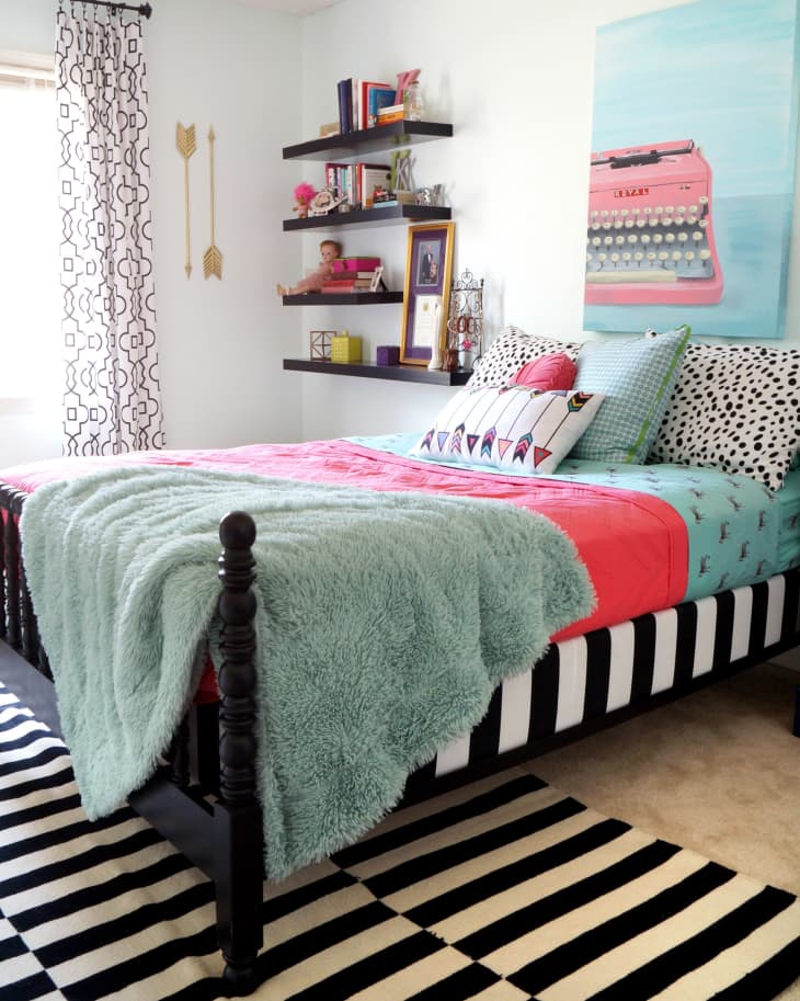 Bedroom with a black bed frame, colorful bedding, striped rug, wall shelves, and a pink typewriter art piece.
