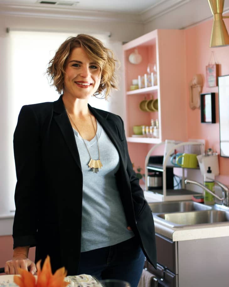 Woman in a black blazer and blue shirt standing in a pink kitchen with open shelves and a stainless steel sink.