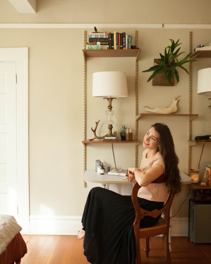 Woman sitting at a small desk in a cozy room with bookshelves, plants, and a lamp.