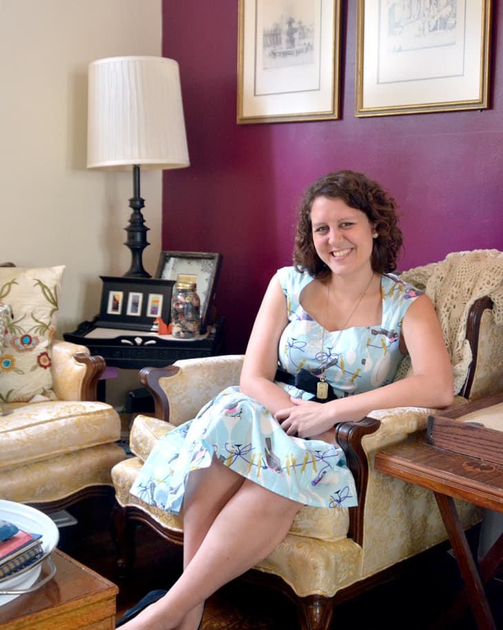 Woman in a floral dress sitting on a vintage armchair in a living room with purple walls and framed art.