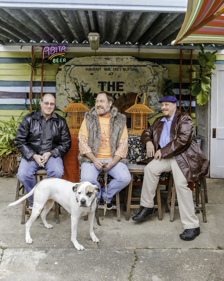 Three men sitting on stools outside a colorful bar with a large dog in front, surrounded by plants and neon signs.