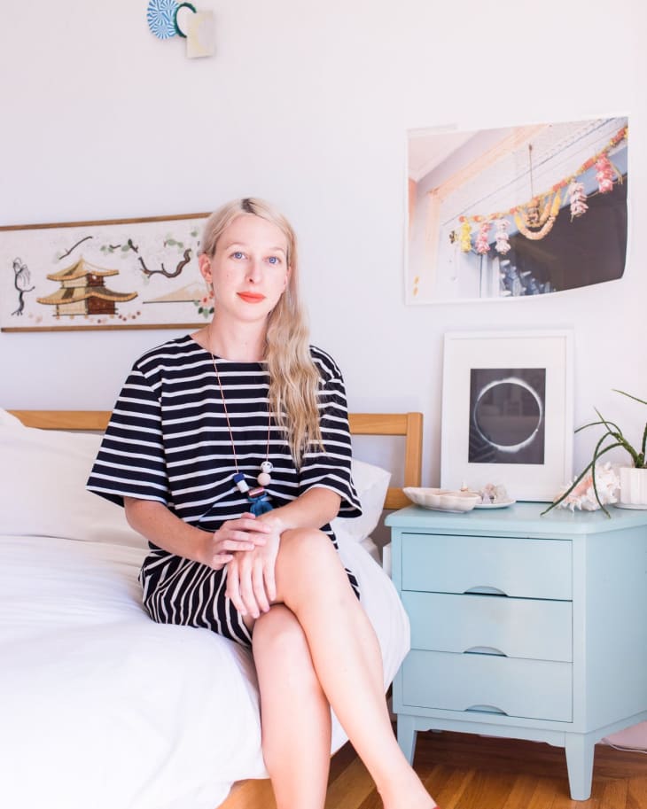 Woman in striped dress sitting on bed, next to a blue nightstand with decor, in a bright room with art on the walls.