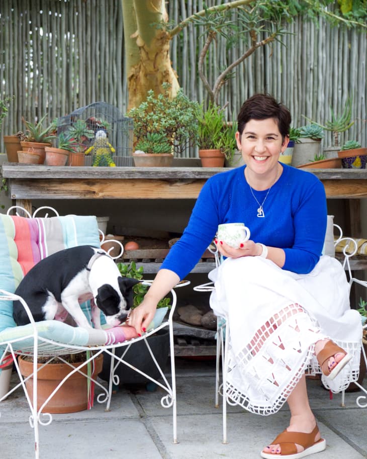 Woman in blue sweater sitting on patio with a small dog, surrounded by potted plants and a wooden table.