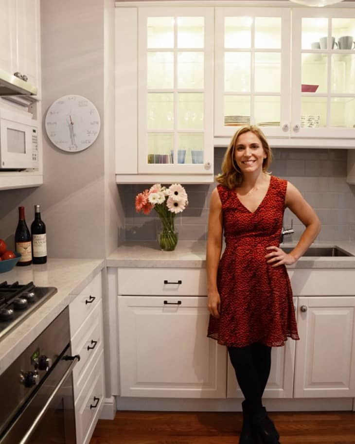 Woman in a red dress standing in a white kitchen with wooden floors, flowers on the counter, and wine bottles.